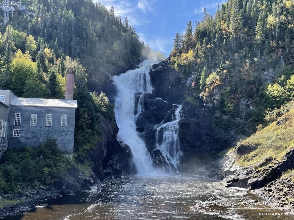 Chute d'eau Ouiatchouan qui dévale une falaise avec un bâtiment gris à sa gauche à Val-Jalbert au Lac Saint-Jean